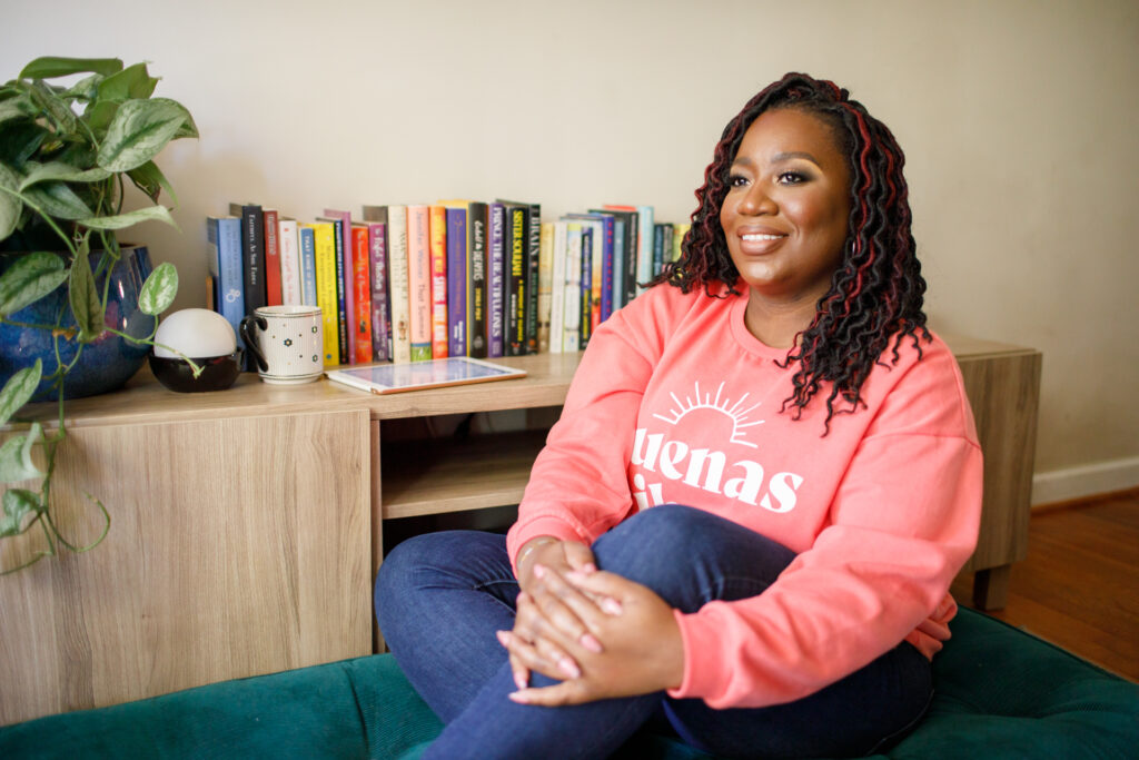 Smiling woman seated by bookshelves wearing pink sweatshirt