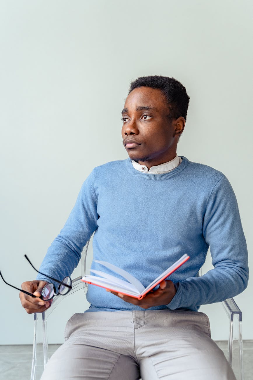 man in blue sweater holding a book and a black framed eyeglasses